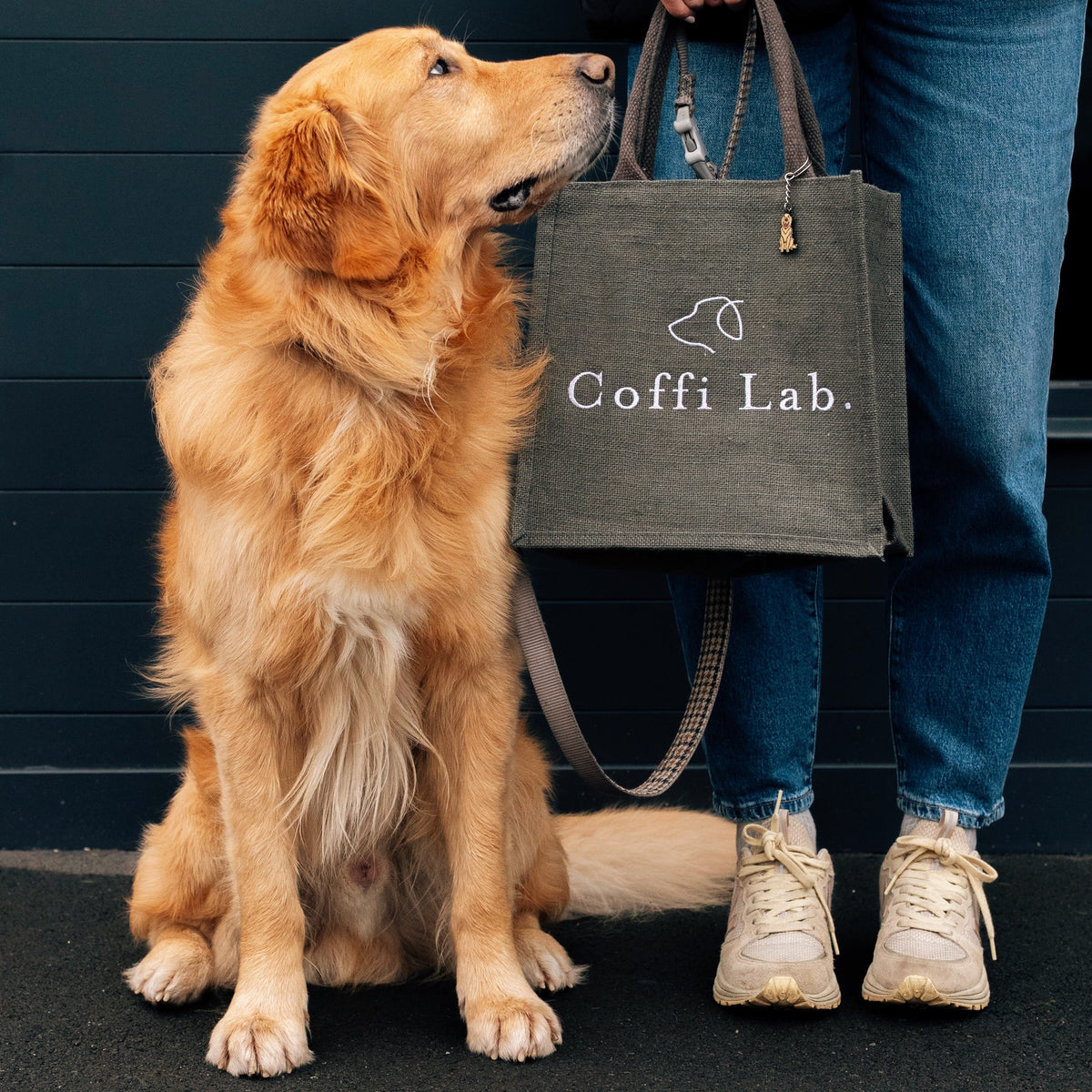 A golden retriever sits next to a person holding a grey tote bag embroidered with a dog-ear logo and the text “Coffi Lab.”