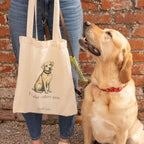 A person holds a 39 x 41cm cream-colored “I Labradore you” Tote - Yellow featuring a Labrador illustration, with a yellow Labrador retriever sitting next to them.